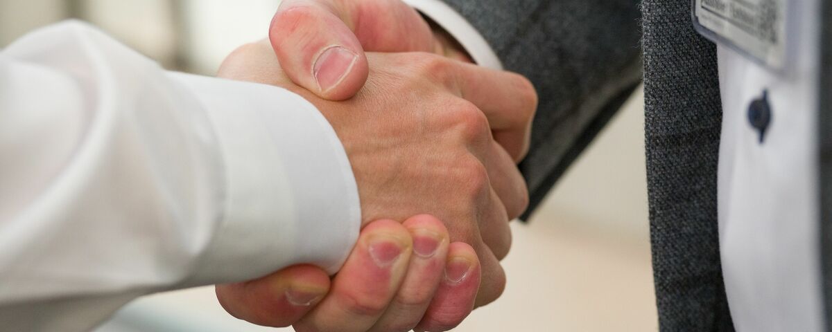 Two businessmen in suits shaking hands in a professional setting - Reynaers