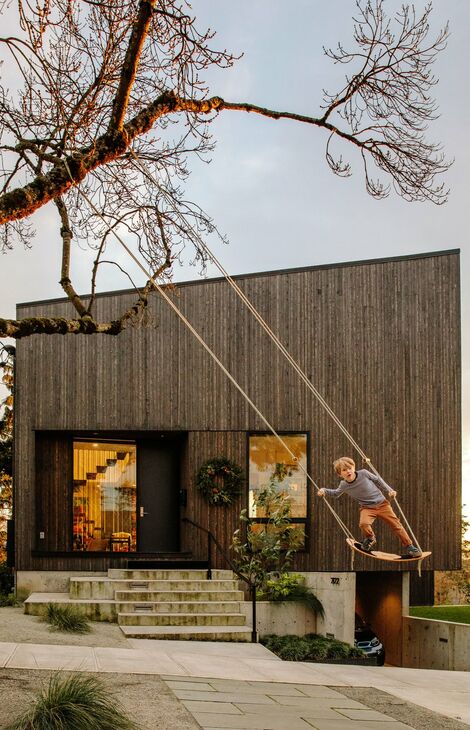 Person swinging on a rope swing attached to a tree in front of a modern wooden house with large windows - Reynaers